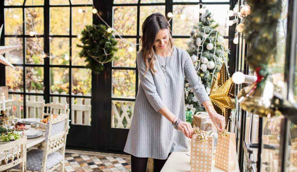 A woman in a stylish grey dress wrapping gifts in a beautifully decorated room with Christmas trees, fairy lights, and festive decorations, with a dining table set for a holiday gathering