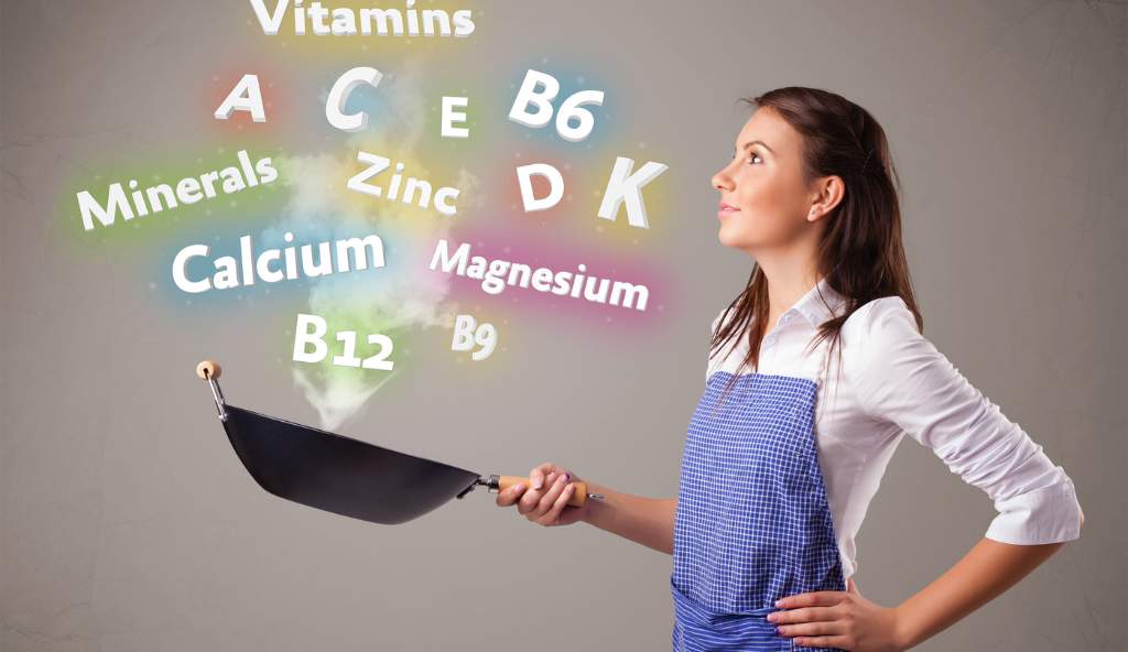 Young woman wearing a blue apron, holding a frying pan with colorful floating text representing vitamins and minerals, symbolizing healthy cooking and nutrition